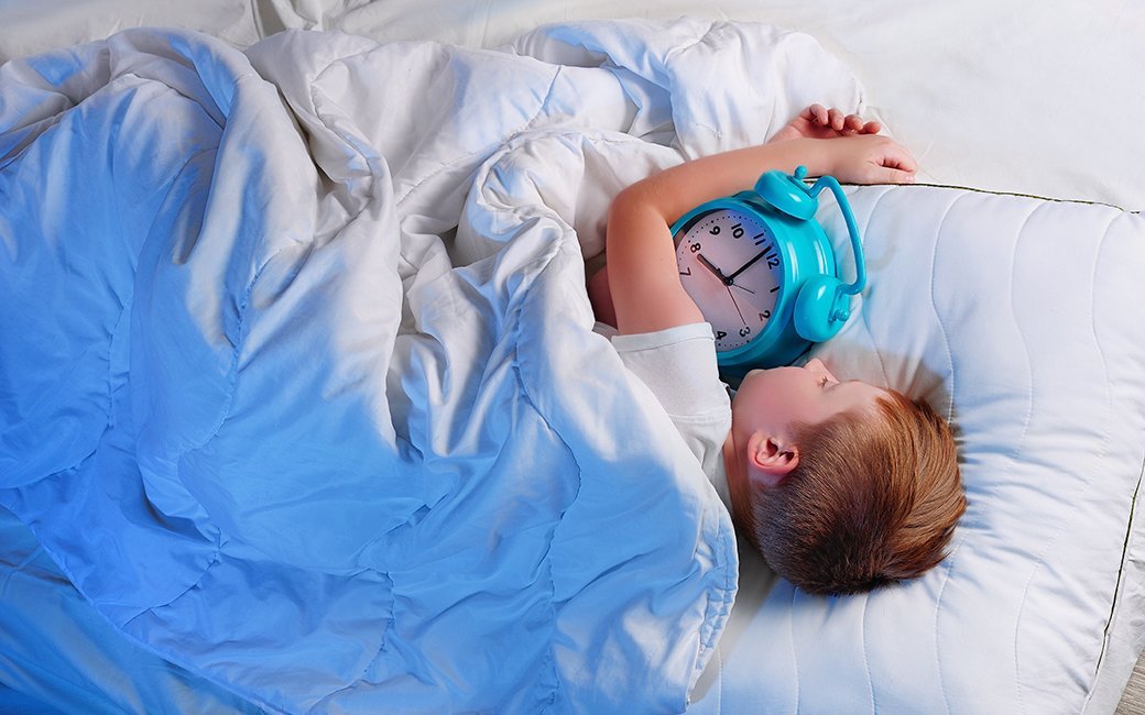 A young child sleeps under a white blanket and holds a large blue alarm clock.