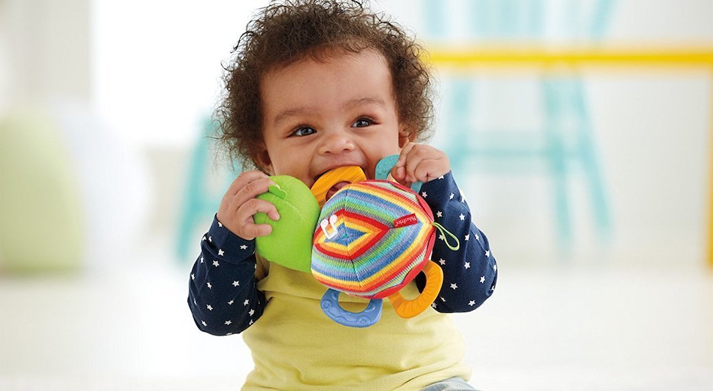 A baby with curly, brown hair chews gleefully on a teething toy.